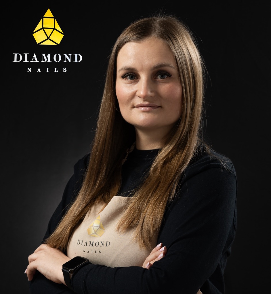 Marina, stylistka Diamond Nails, z założonymi rękami, w beżowym fartuchu z logo salonu, na czarnym tle